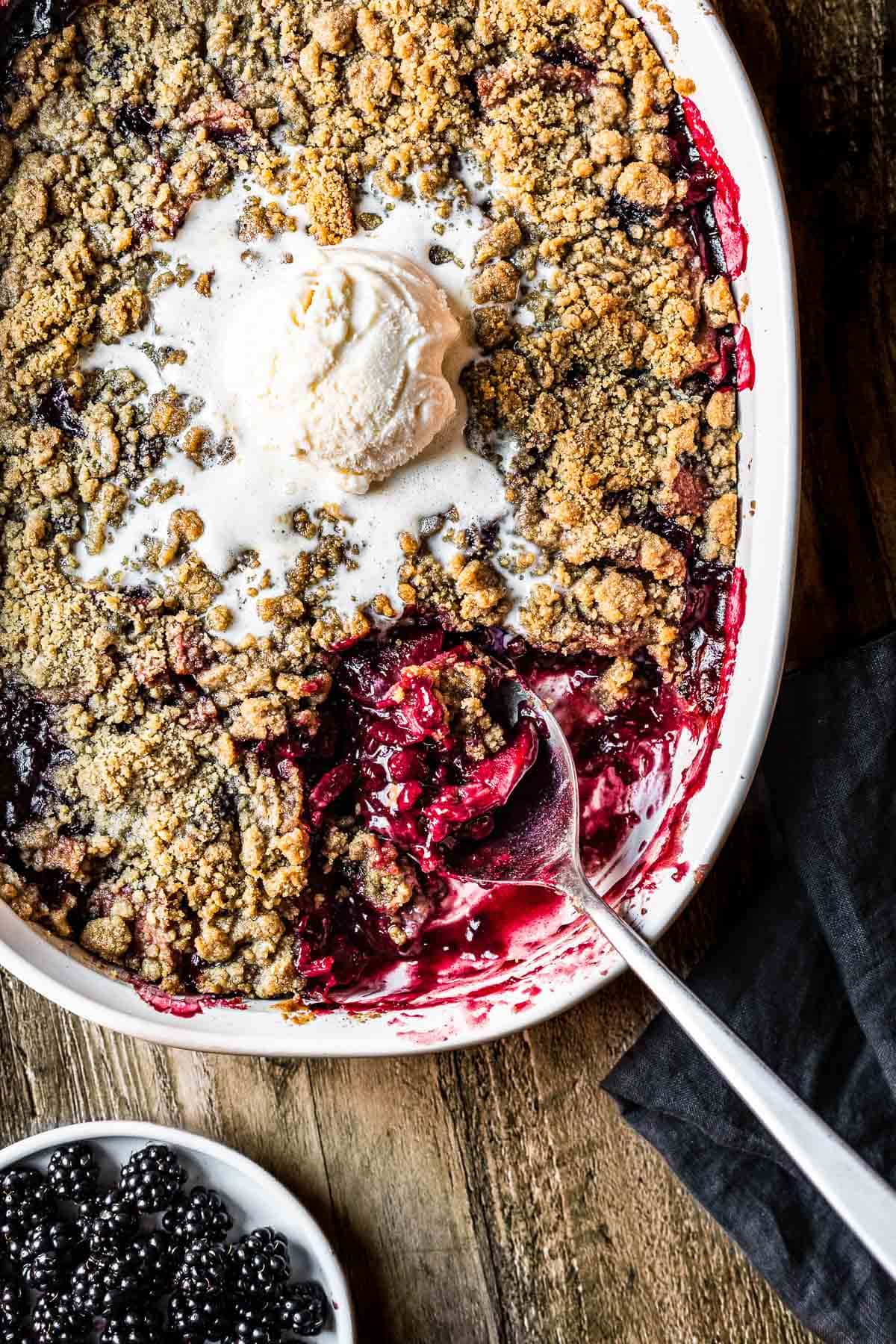 Apple and berry crumble in a white ceramic dish with a spoon and a scoop of vanilla ice cream on top. The table underneath is rustic wood.