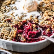 A spoon scoops fruit crumble from a casserole dish with a scoop of vanilla ice cream on top.