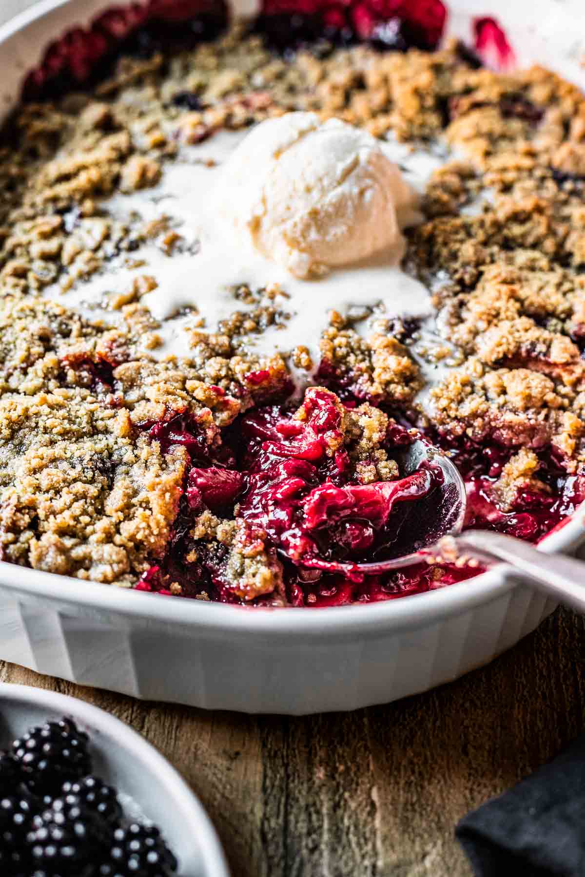 A spoon scoops fruit crumble from a casserole dish with a scoop of vanilla ice cream on top.