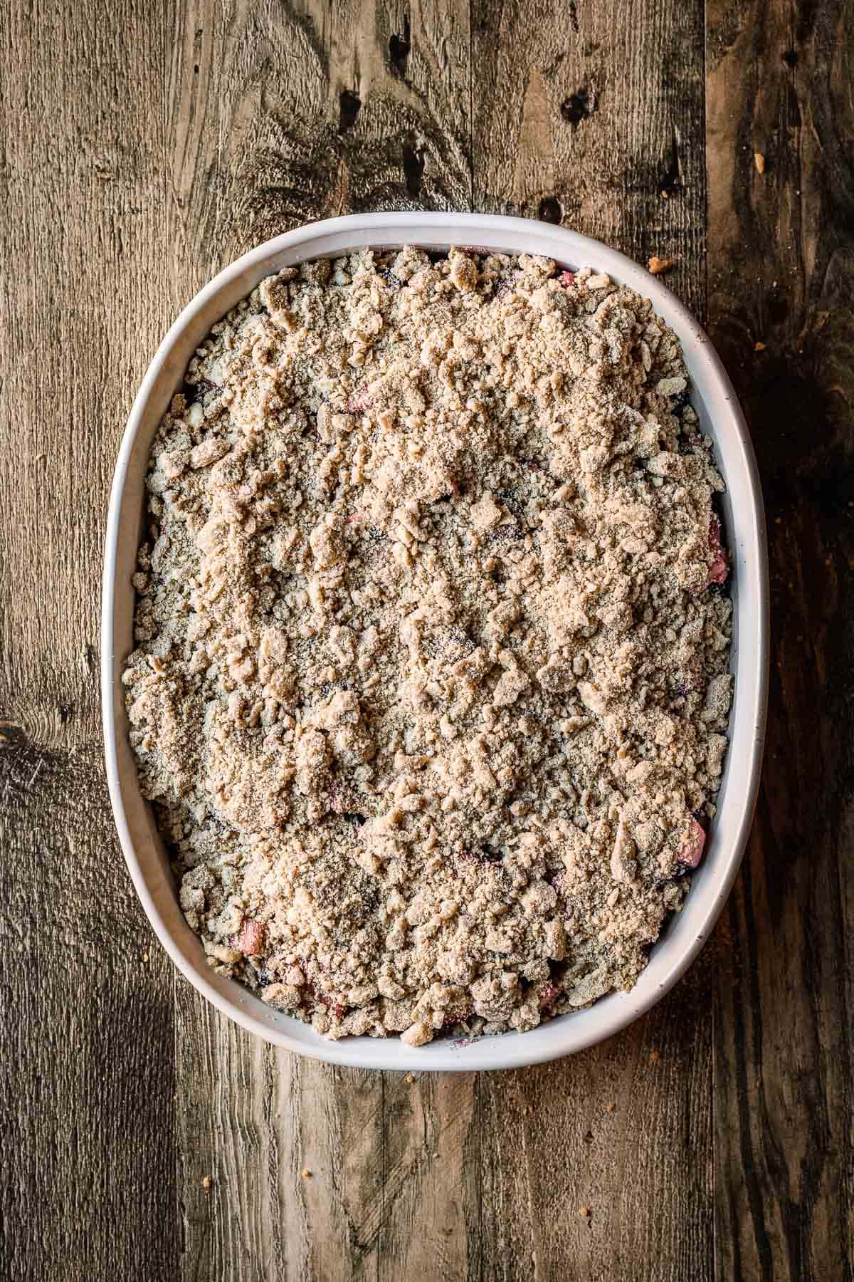 A white ceramic baking dish filled with fruit and covered with crumble topping on a rustic wood tabletop.