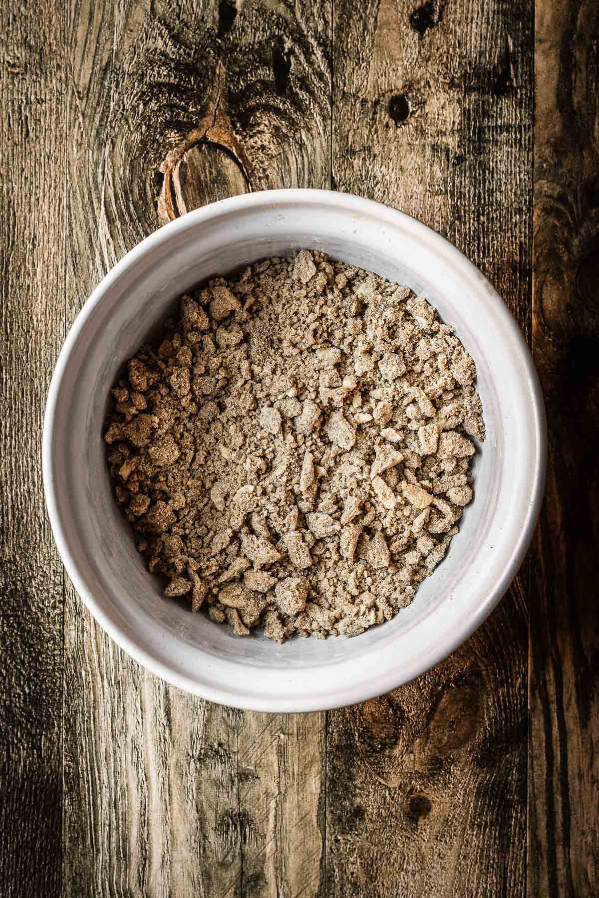 A white bowl filled with crumble topping for a fruit crumble. The bowl rests on a rustic wooden tabletop.