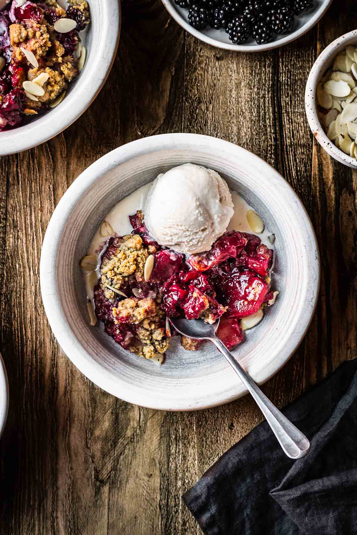 A serving of apple and blackberry crumble in a white ceramic bowl with a spoon and a scoop of ice cream on the side. The bowl rests on a rustic wooden table.