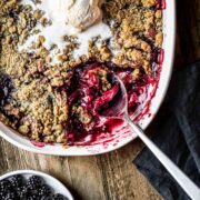 Blackberry and apple crumble being scooped out of a white ceramic dish with a scoop of vanilla ice cream on top.