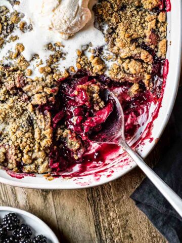 Blackberry and apple crumble being scooped out of a white ceramic dish with a scoop of vanilla ice cream on top.