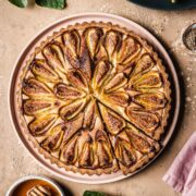 A baked tarte aux figues with a slice cut out, with fig leaves and honey nearby.