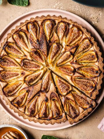 A baked tarte aux figues with a slice cut out, with fig leaves and honey nearby.