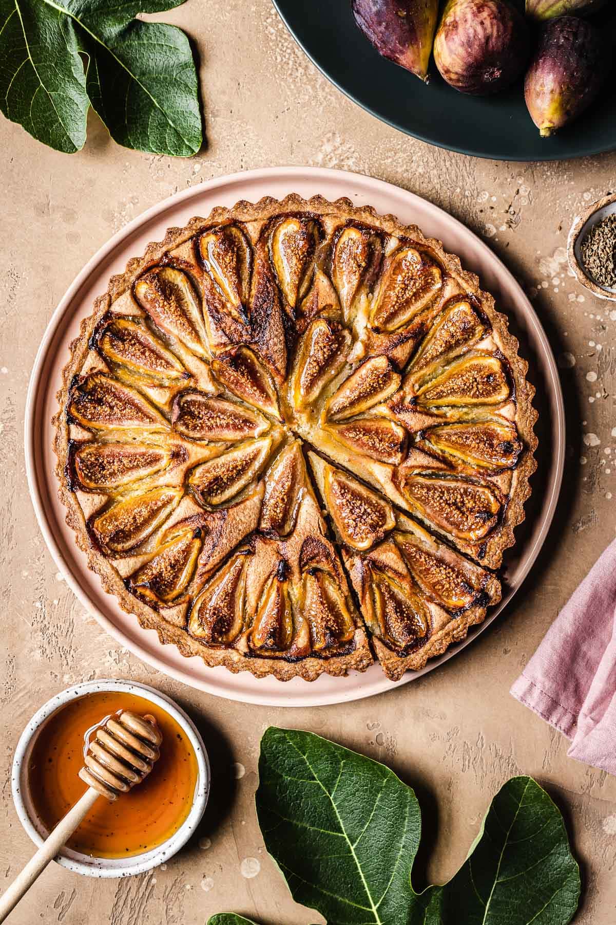 A baked tarte aux figues with a slice cut out, with fig leaves and honey nearby.