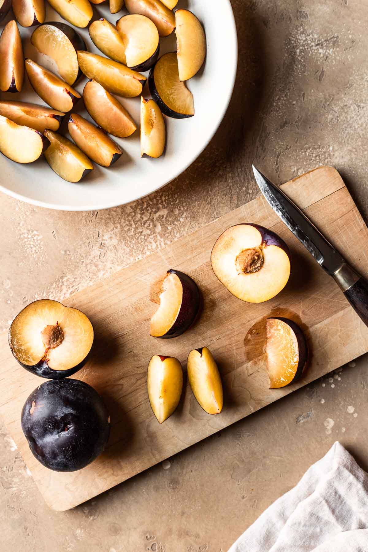 Sliced plums on a wooden cutting board with more on a plate nearby.