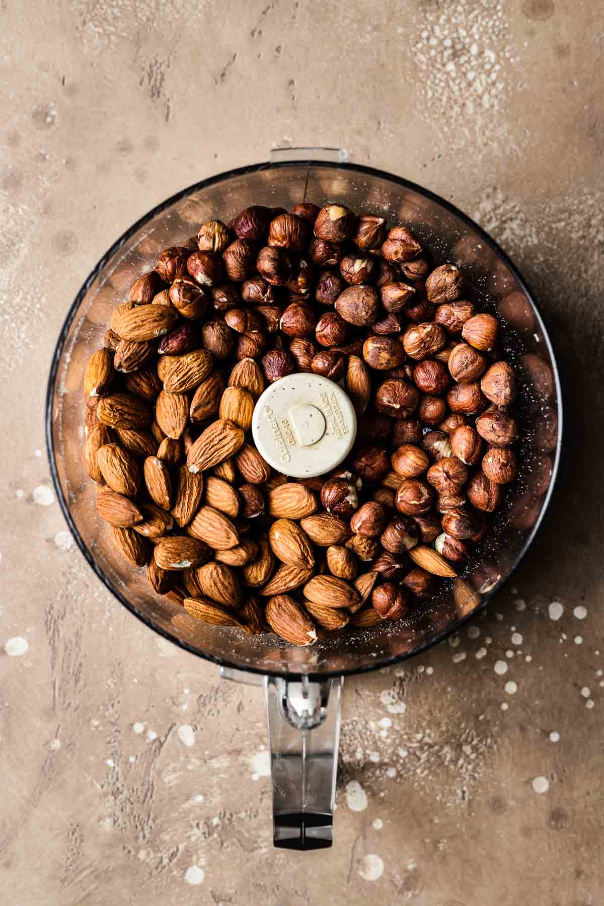 A food processor bowl filled with half almonds and half hazelnuts.