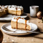 A slice of gingerbread cheesecake topped with whipped cream and caramel sauce on a white plate.