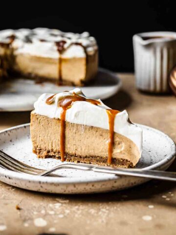 A slice of gingerbread cheesecake topped with whipped cream and caramel sauce on a white plate.