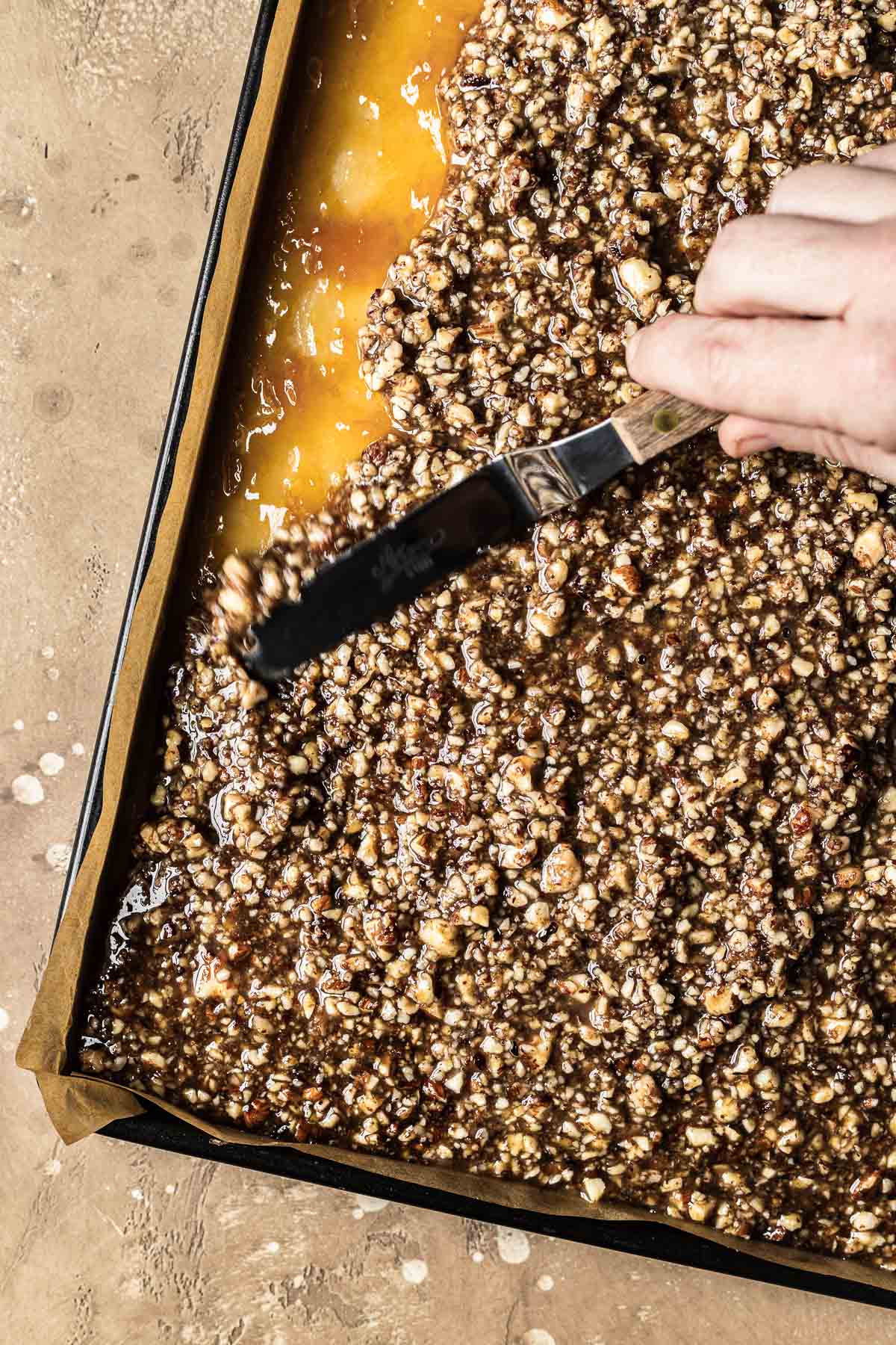 A hand holding a mini spatula spreads nussecken nut topping over apricot jam.