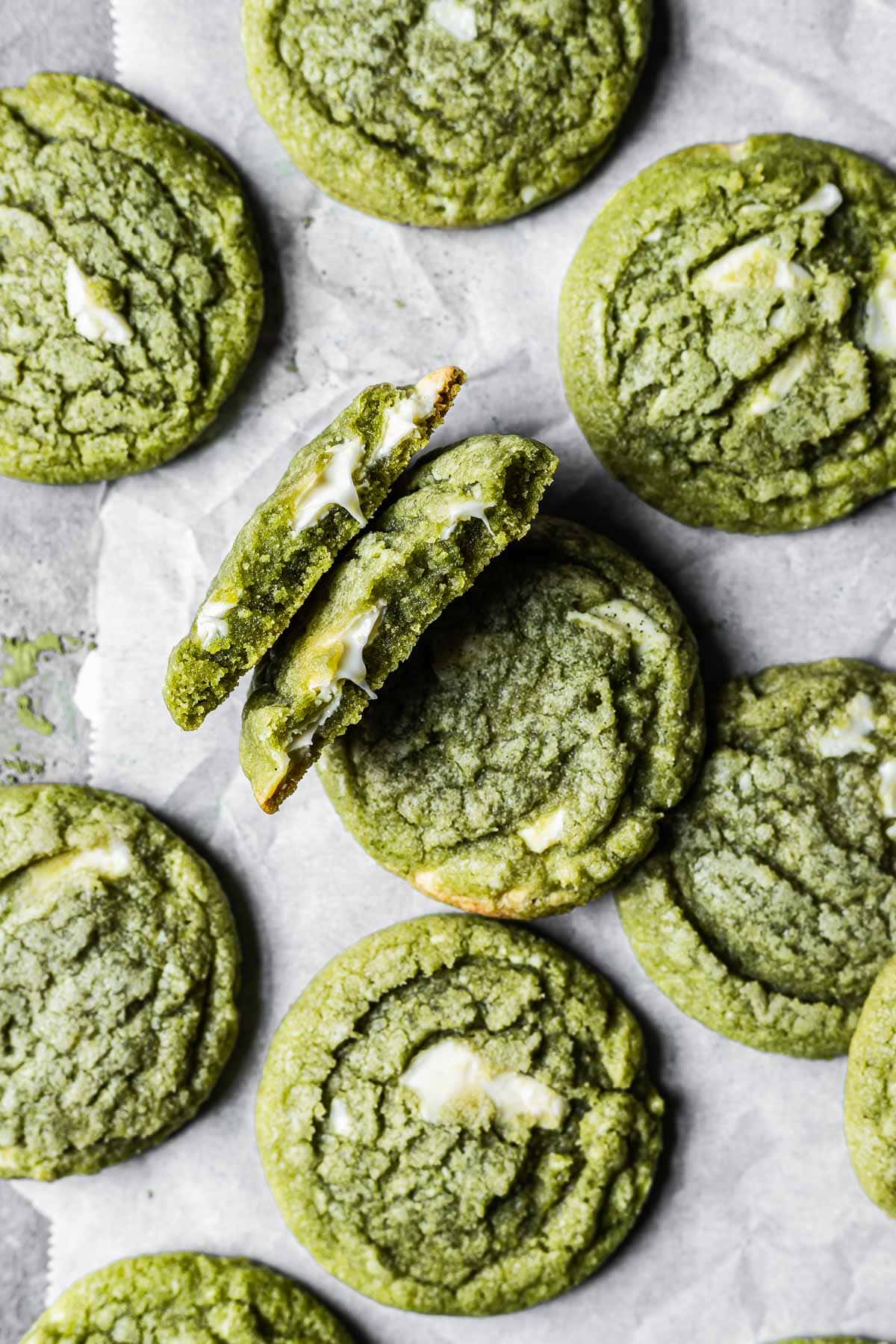 A matcha white chocolate cookie is broken in half, revealing the gooey melted chocolate inside. There are many other matcha cookies surrounding it.