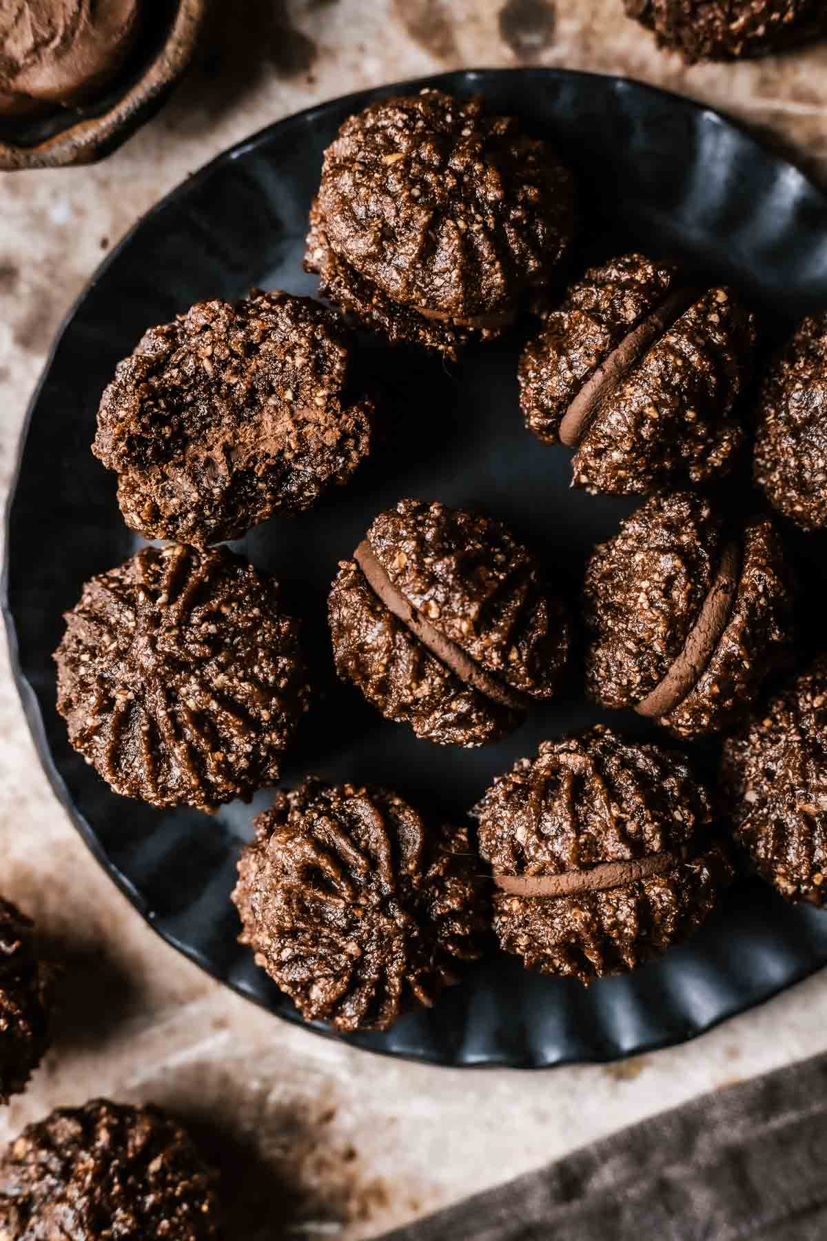 Piped chocolate hazelnut sandwich cookies filled with chocolate ganache on a black ceramic plate.