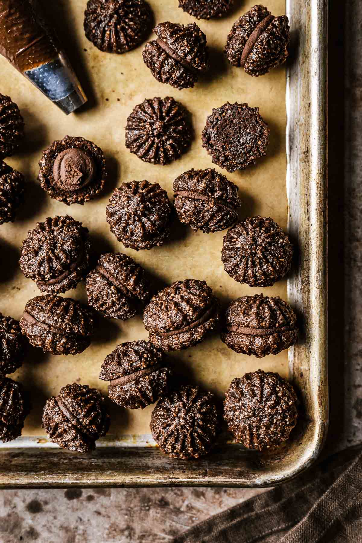 A piping bag of chocolate ganache on a baking sheet with chocolate hazelnut sandwich cookies.