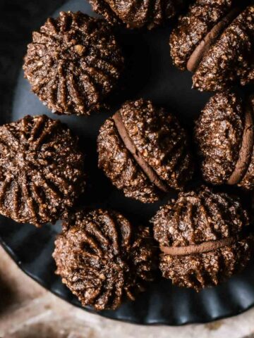 Close up view of piped chocolate hazelnut sandwich cookies filled with chocolate ganache on a black ceramic plate.