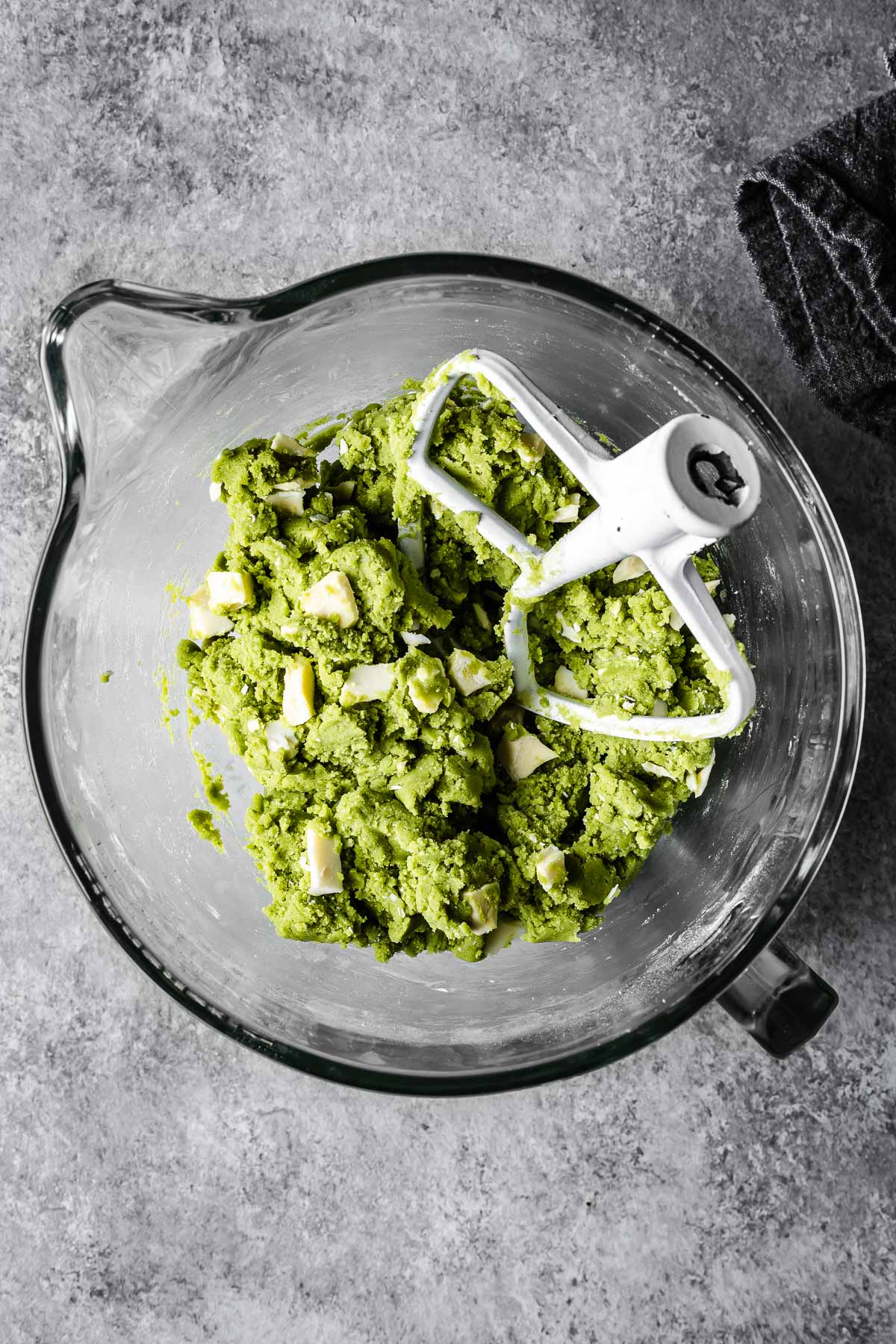 Green cookie dough in a glass mixer with white chocolate chunks added in.