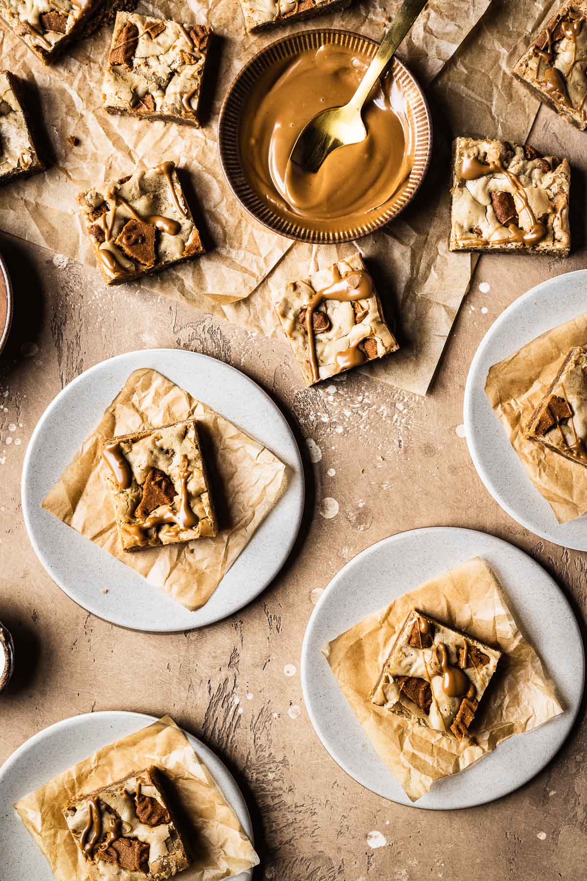 Plates of sliced bar cookies on brown parchment squares with a small bowl of biscoff spread nearby.