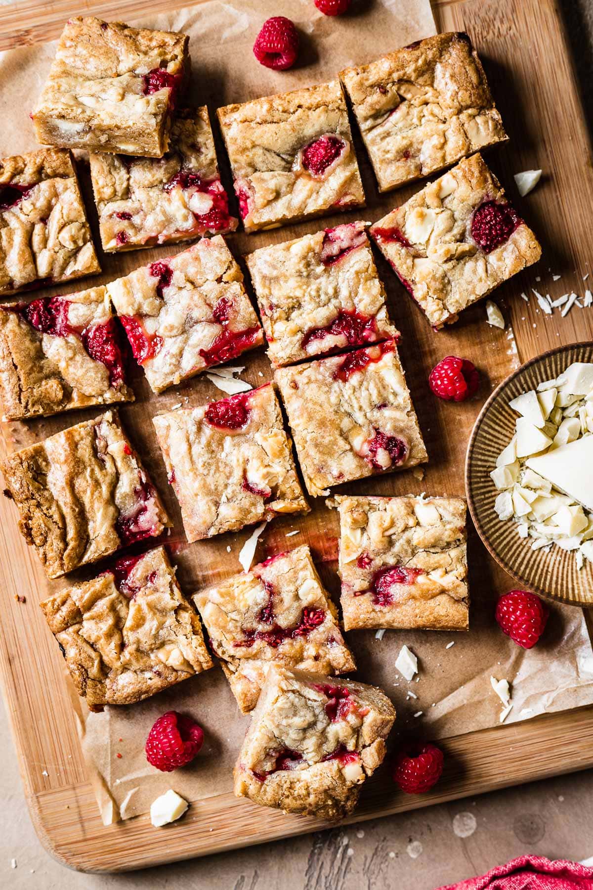 Raspberry white chocolate blondie squares on a wooden cutting board with a small bowl of chopped white chocolate nearby.