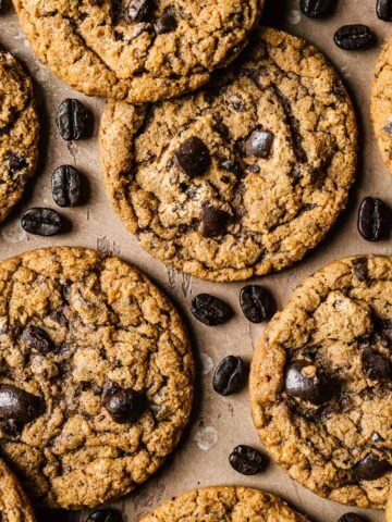 Coffee cookies on a tan surface with coffee beans sprinkled amongst them.