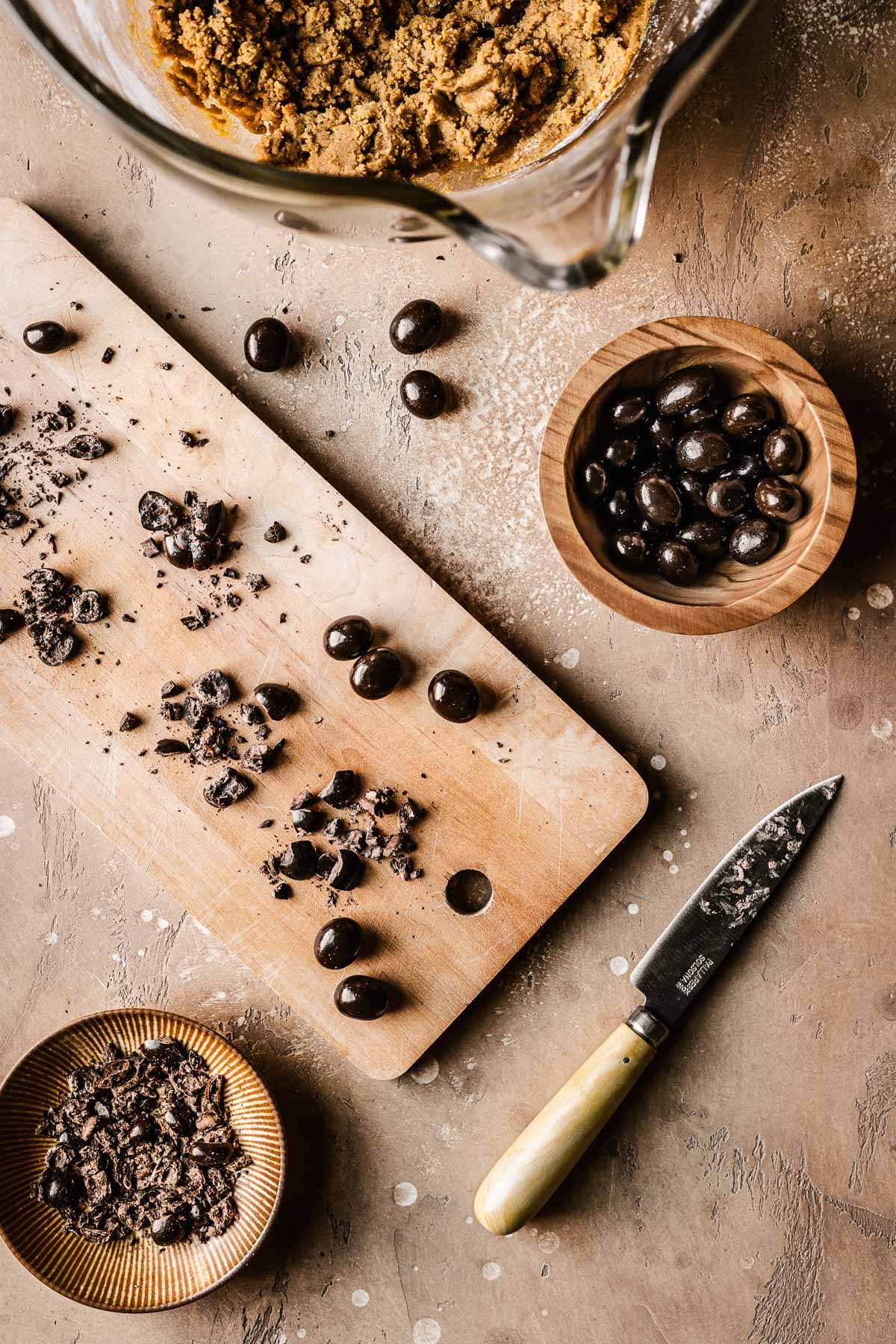 A cutting board with chopped chocolate covered coffee beans, a bowl of whole chocolate coffee beans, and a paring knife on a brown stone surface.