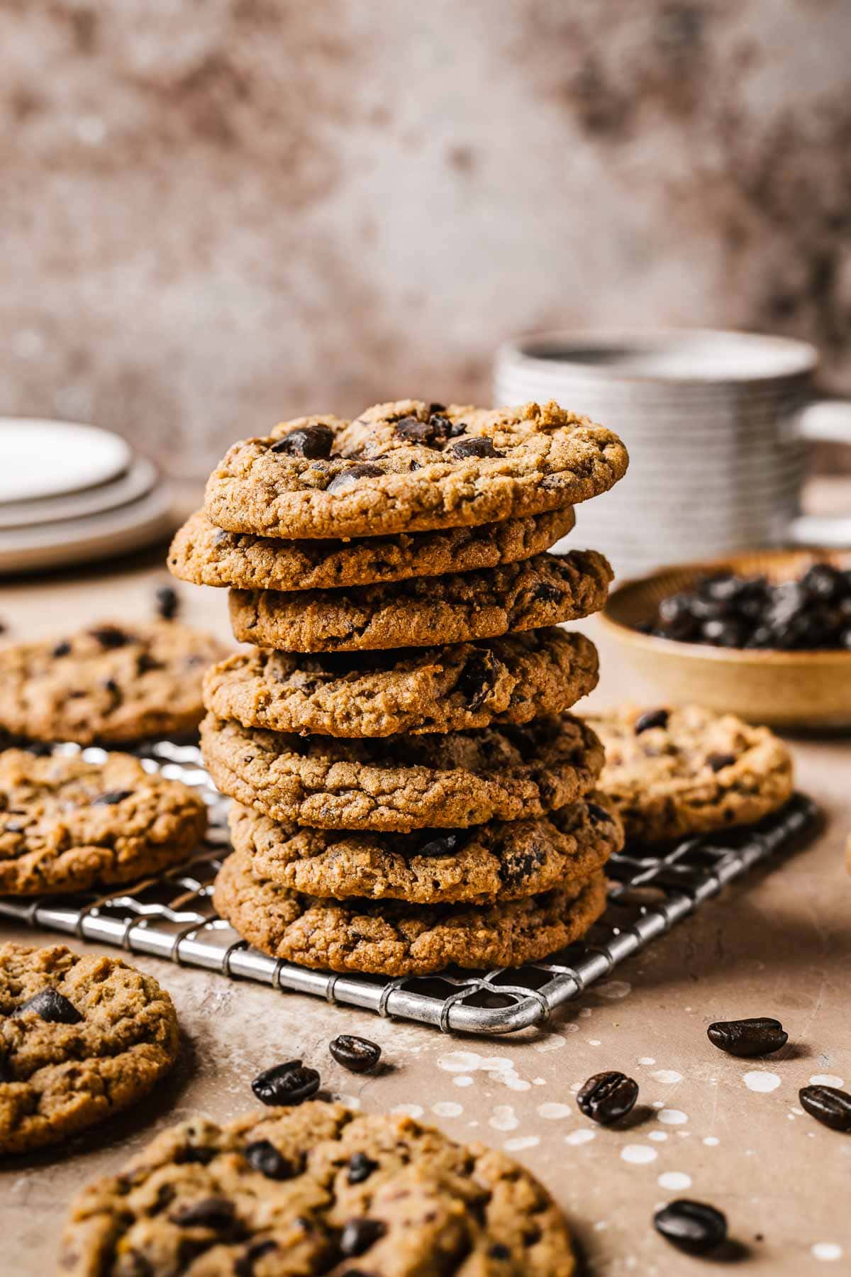 A stack of seven brown cookies on a metal cooling rack with coffee beans in a small bowl and scattered nearby.