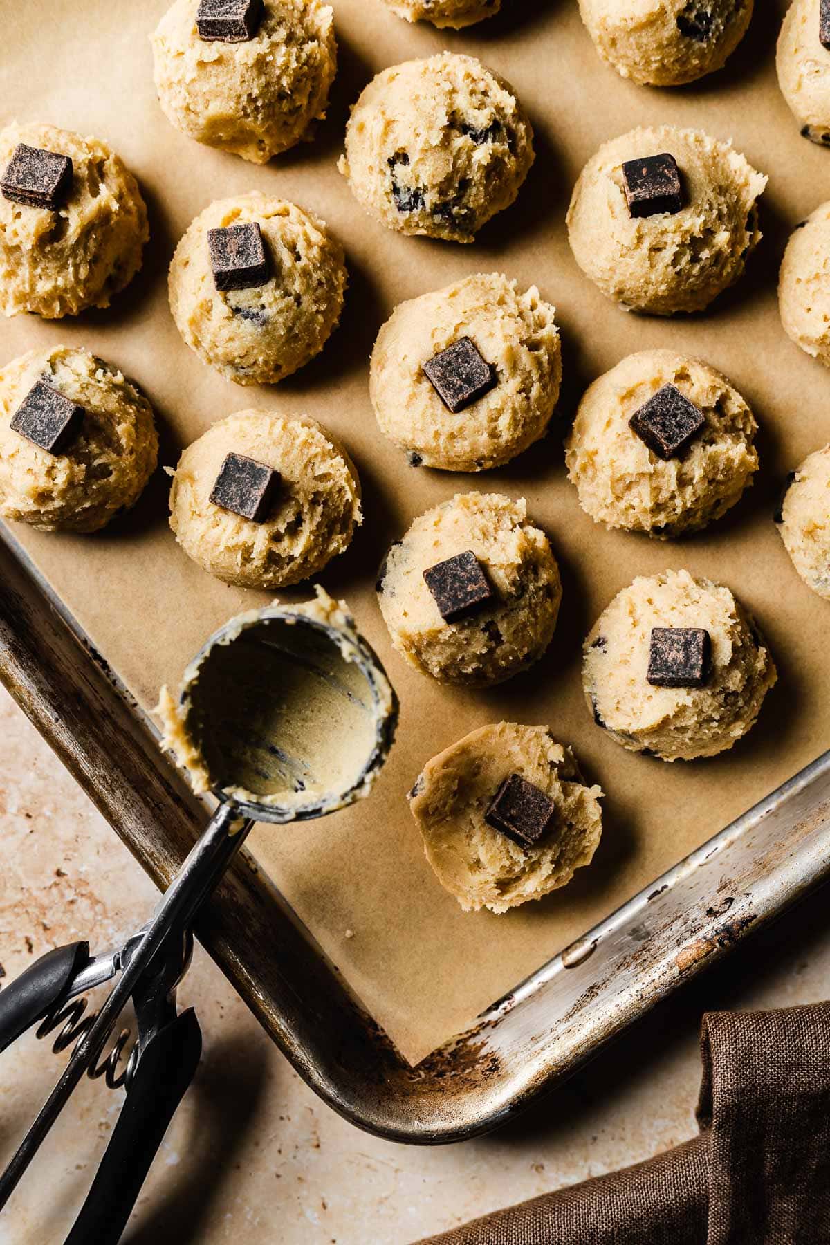 A sheet pan of unbaked cookie dough scoops lined with brown parchment paper.