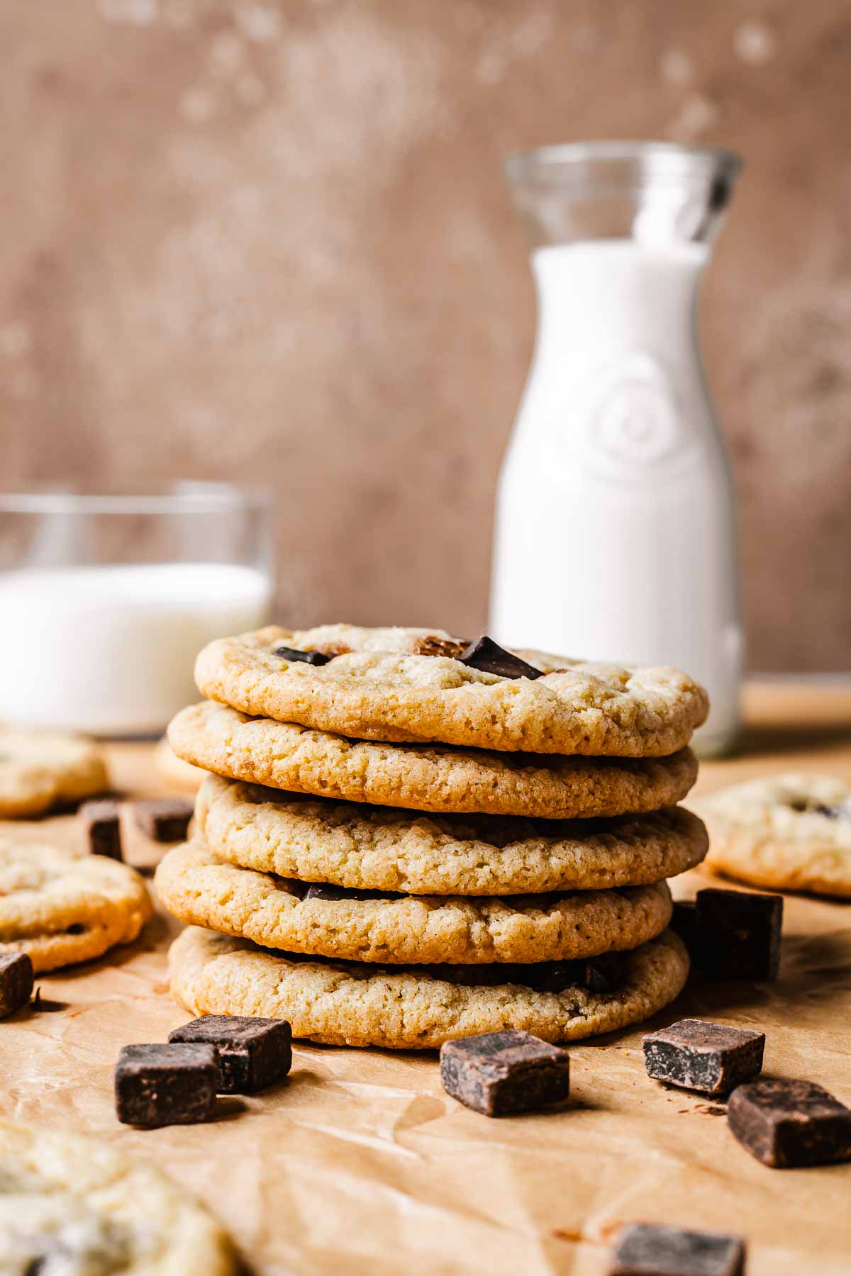A stack of five chocolate chip cookies with a pitcher and glass of milk in the background.
