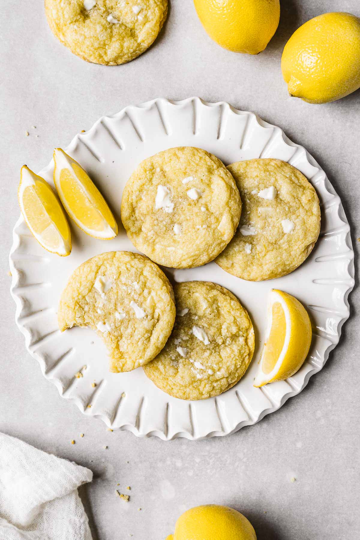 Four lemon white chocolate cookies on a white fluted ceramic plate surrounded by slices of lemons, whole lemons, and another cookie.