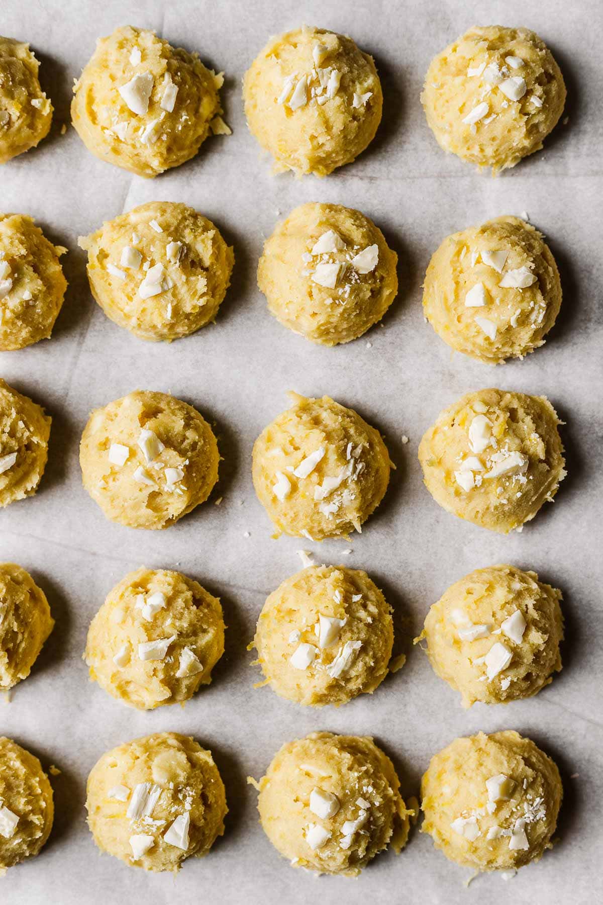Scoops of yellow cookie dough topped with white chocolate chunks on a white parchment lined baking sheet.