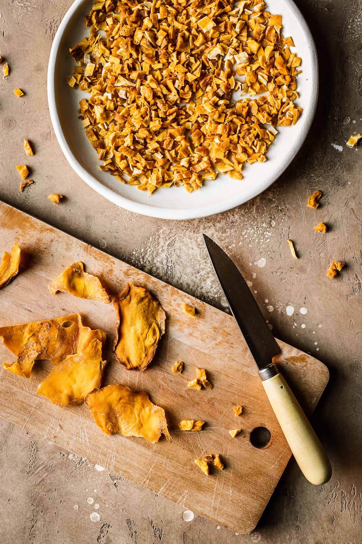 A plate of diced dried mangos alongside a wooden cutting board, paring knife and dried mango cheeks.