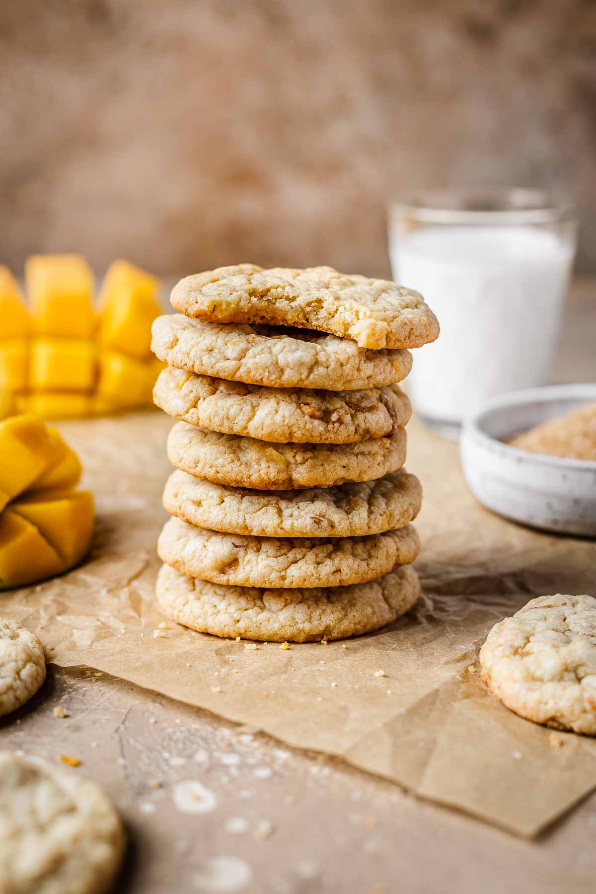 A stack of seven mango cookies on brown parchment paper with a glass of milk and sliced mangos in the background.