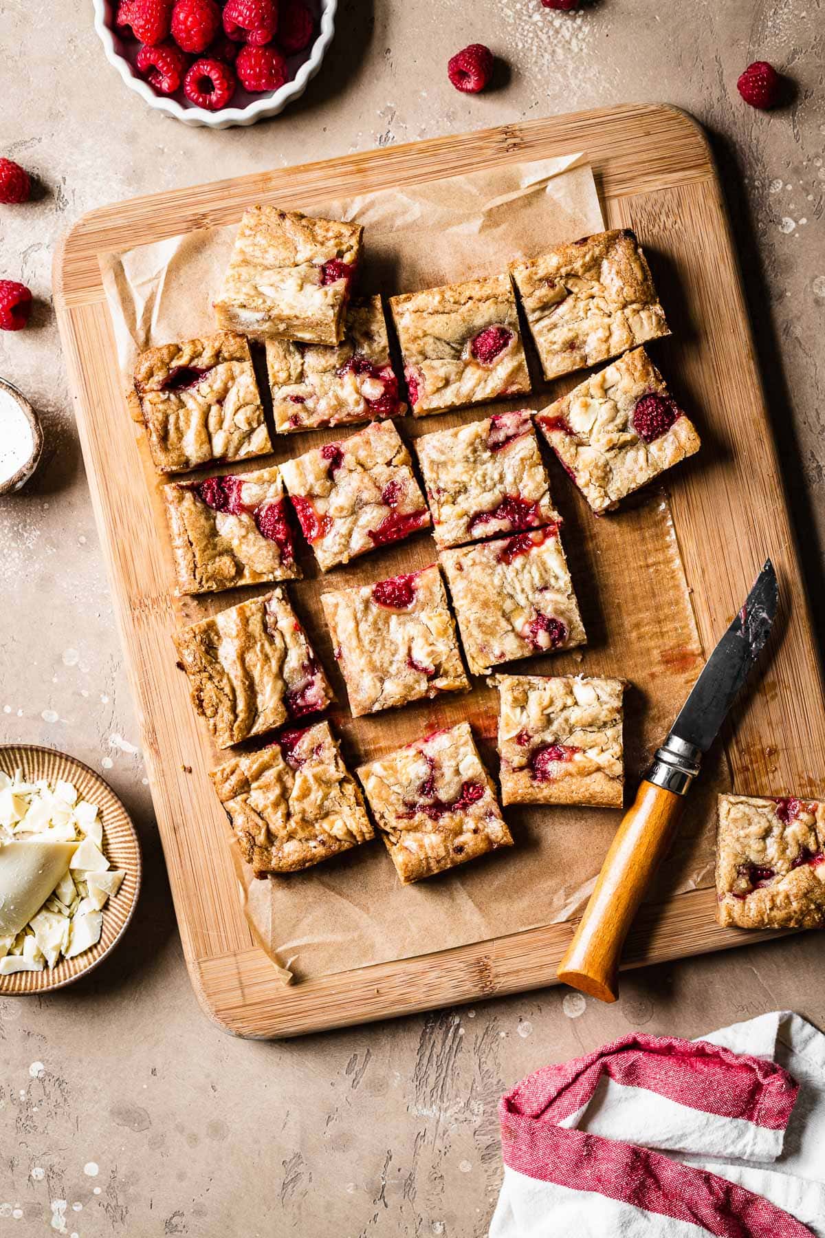 Raspberry and white chocolate blondies cut into squares on a wooden cutting board, surrounded by ingredients, a knife and a white and red napkin.