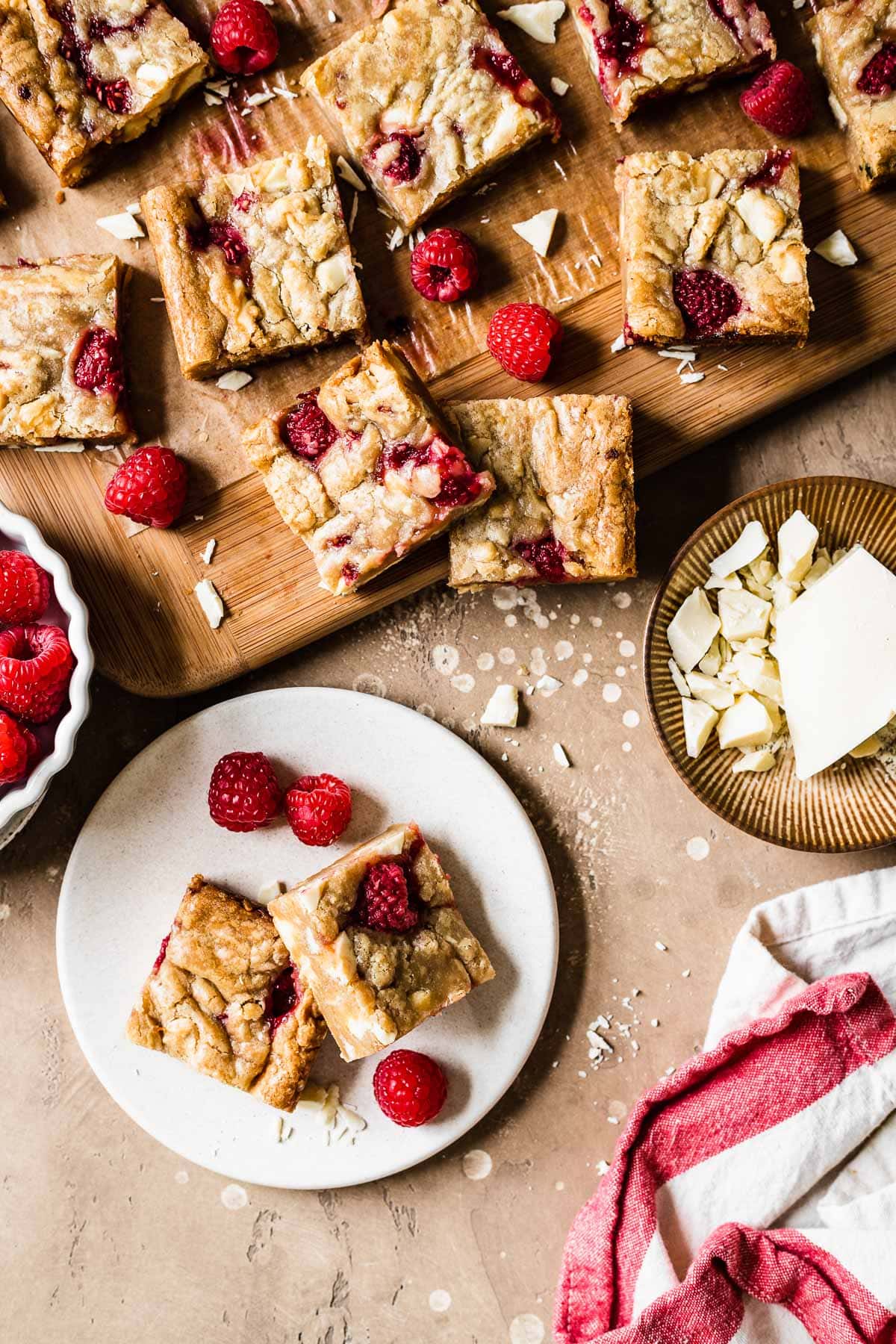 Two blondie bars on a small plate with raspberries. There are more cut blondies on a wooden cutting board nearby, along with a small bowl of chopped white chocolate.