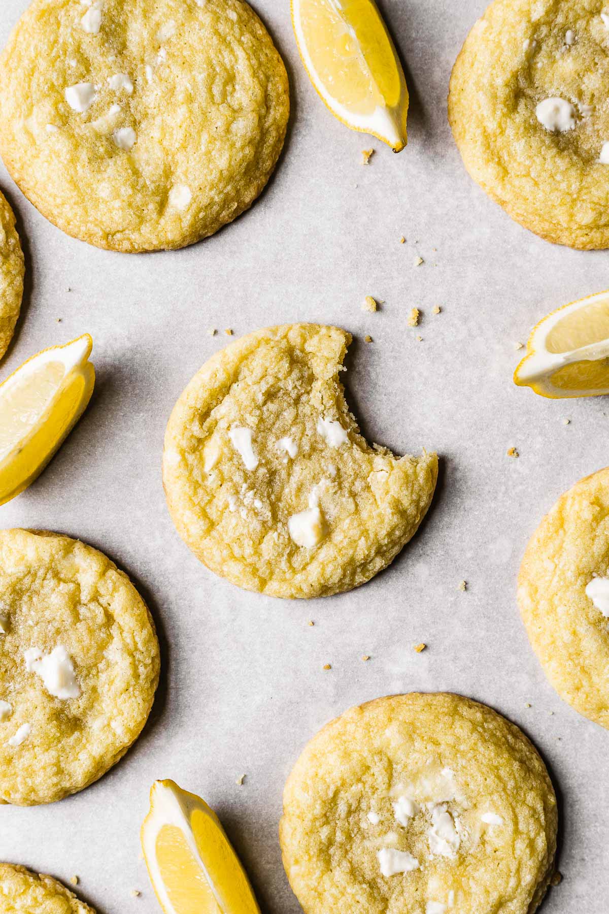 A lemon cookie with a bite taken out of it is surrounded by more cookies and lemon wedges on white parchment paper.