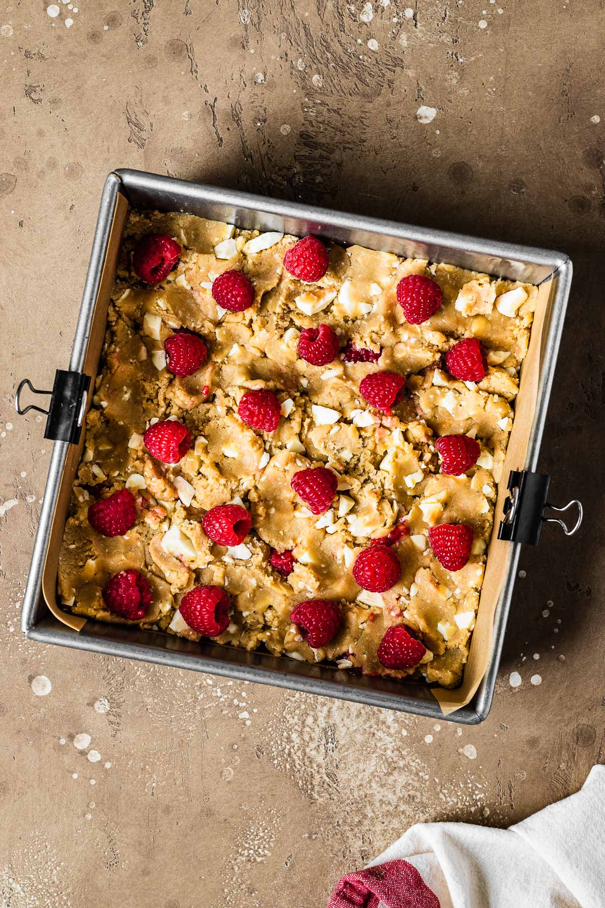 A square parchment lined pan filled with unbaked blondie dough studded with white chocolate and raspberries.