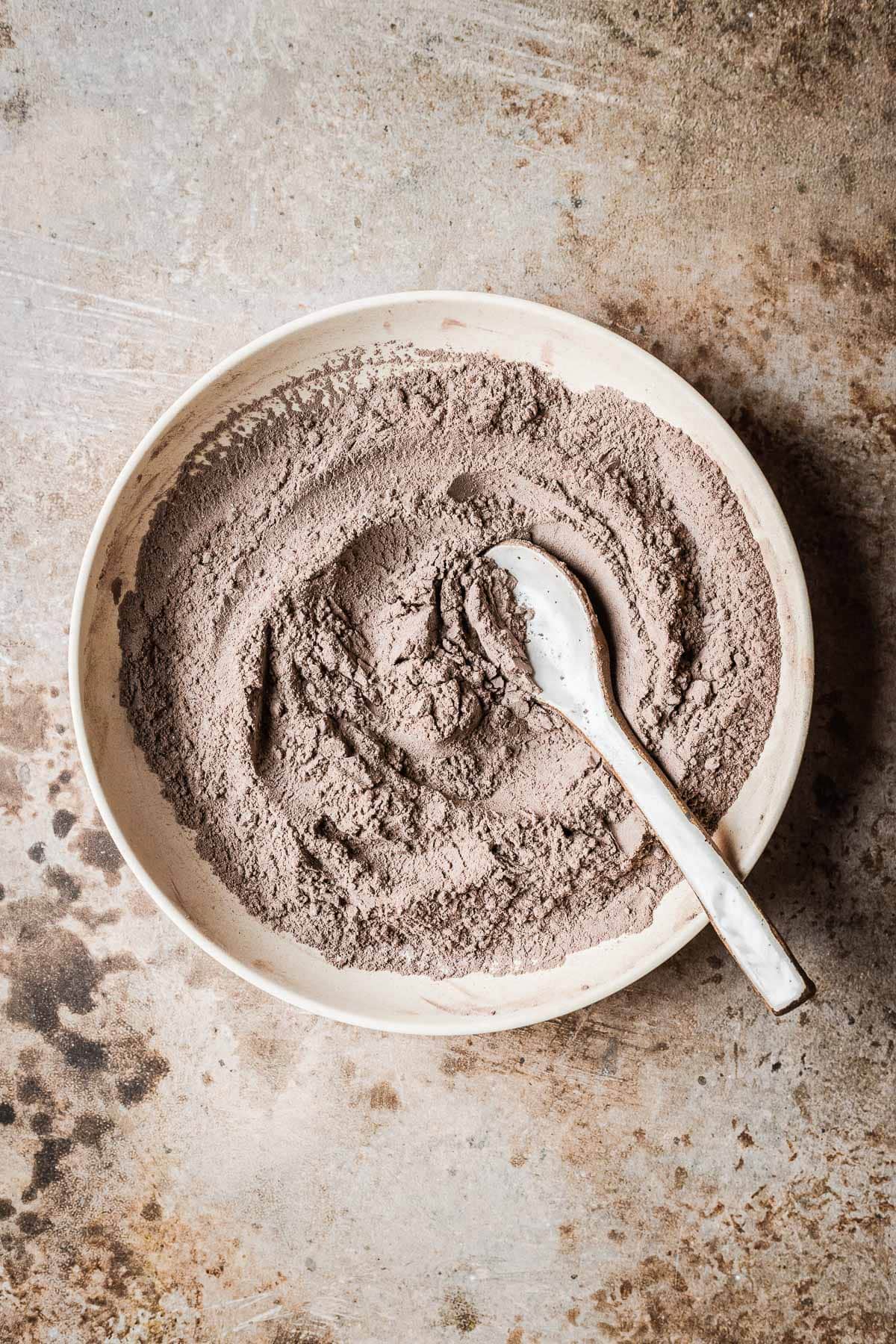 Sifted and mixed ingredients for chocolate cookies with cocoa powder in a tan bowl with a white ceramic spoon.