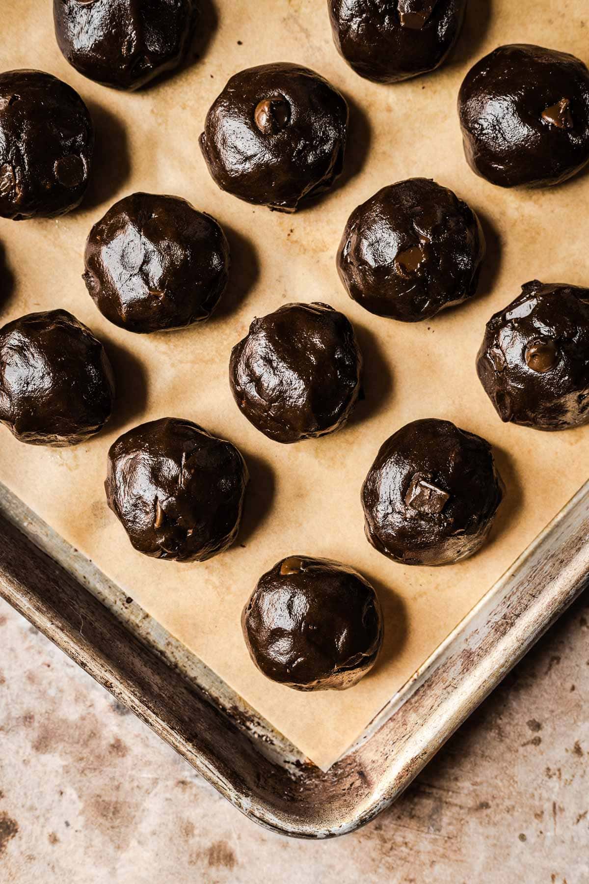 Rolled balls of chocolate cookie dough on a brown parchment lined baking sheet.