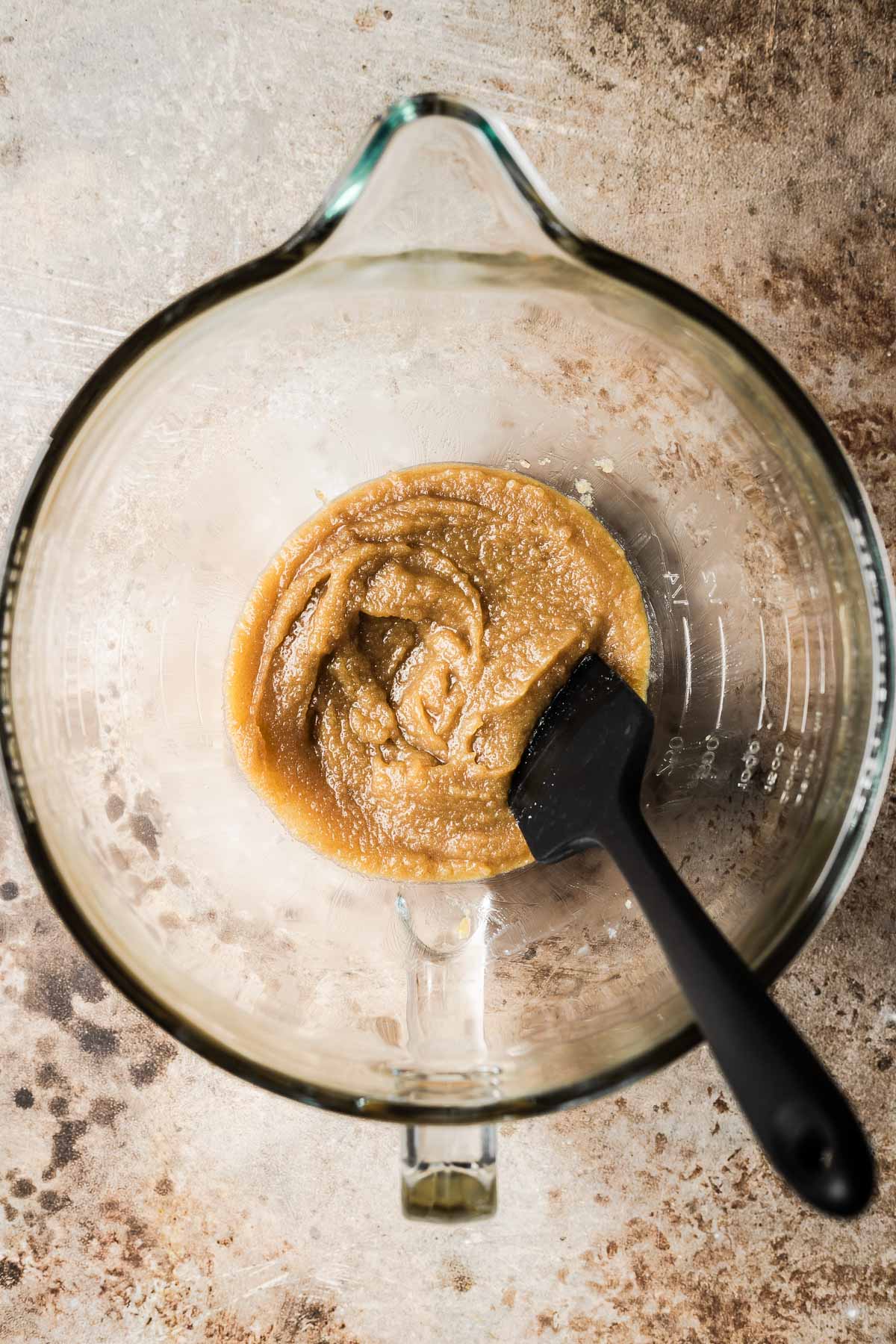 A clear glass mixing bowl with a black spatula mixing melted butter and sugar together.