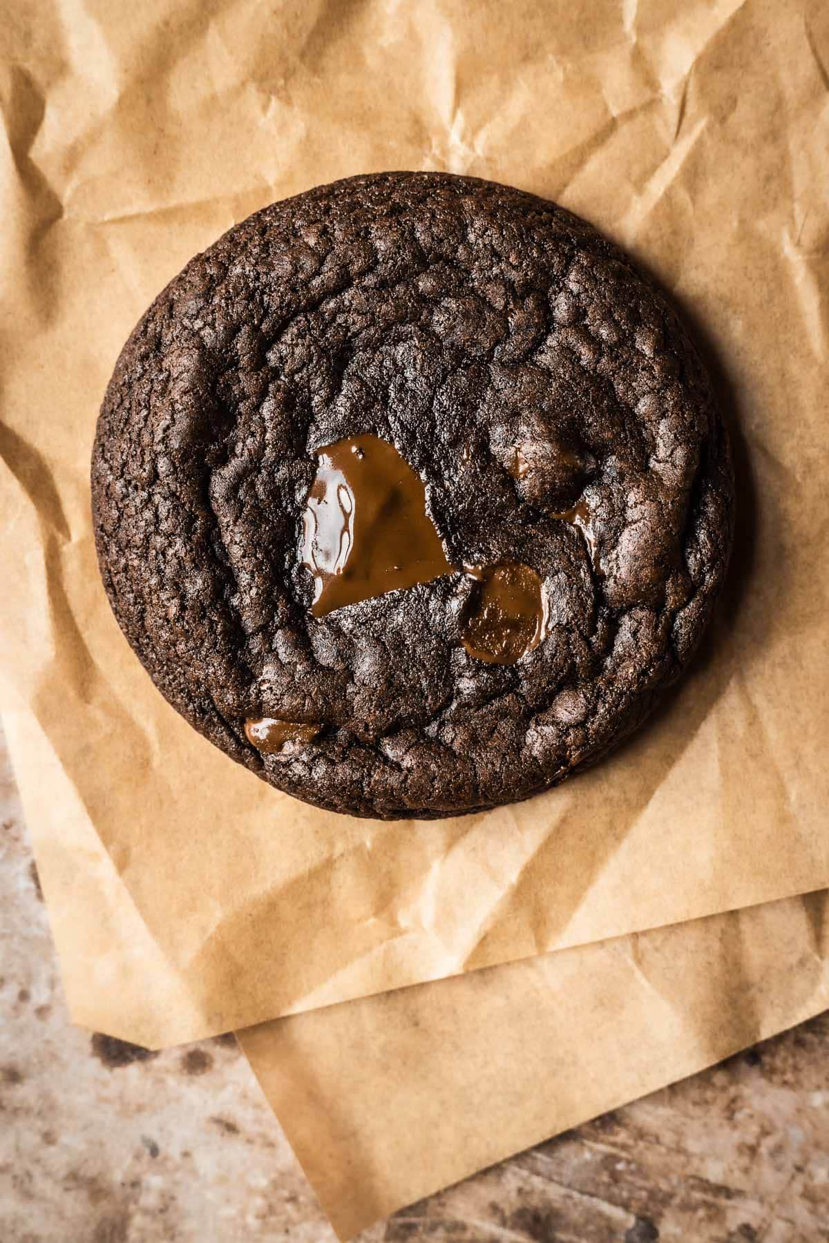 A single double chocolate cookie on brown parchment paper with melted pools of melted chocolate chunks on top.