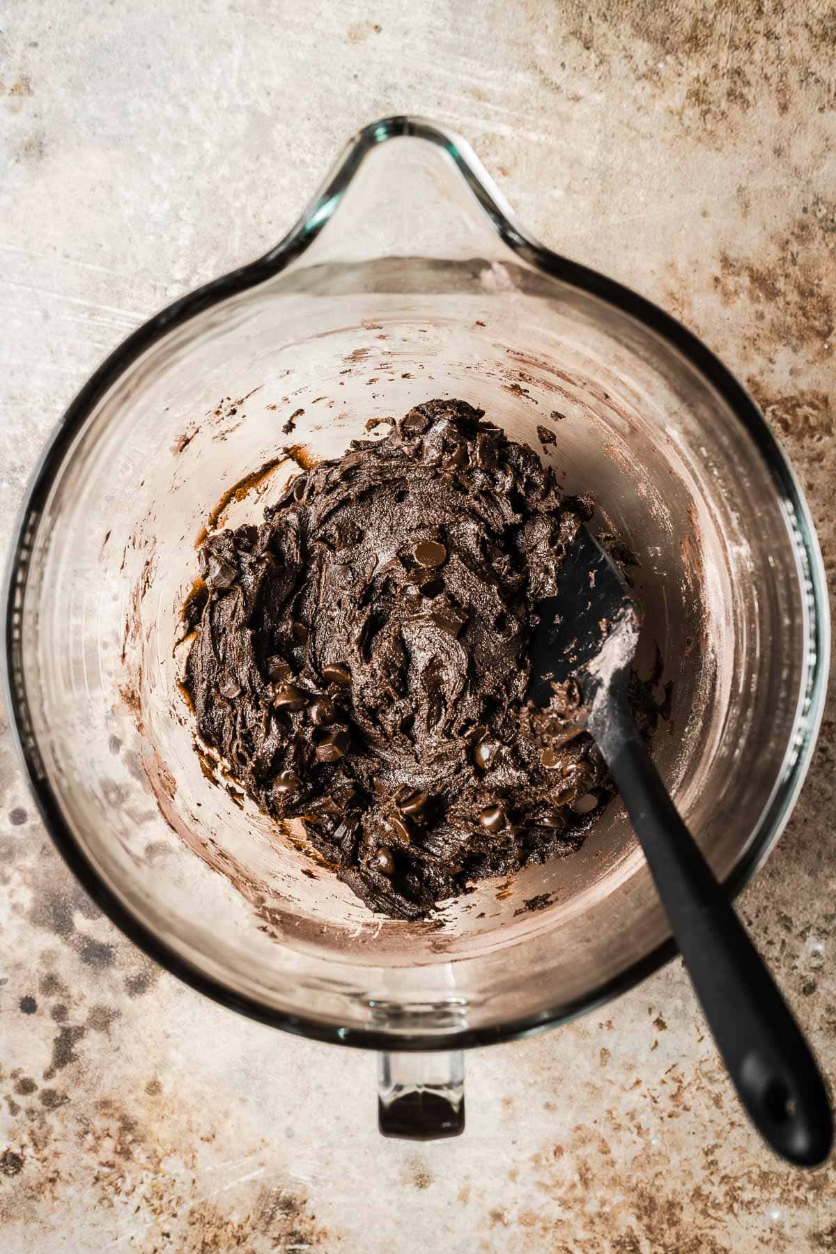 A clear mixing bowl of chocolate cookie dough with chocolate chips and chunks added.