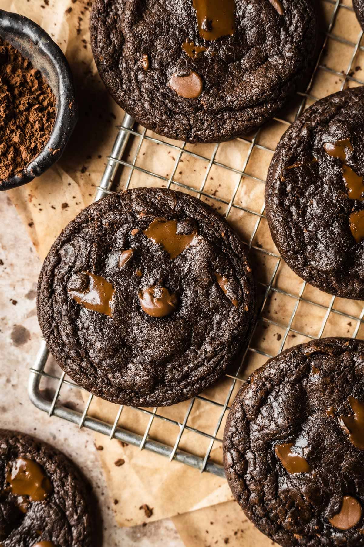 A metal cooling rack with dark chocolate cookies on top of brown parchment paper with a small bowl of cocoa powder nearby.