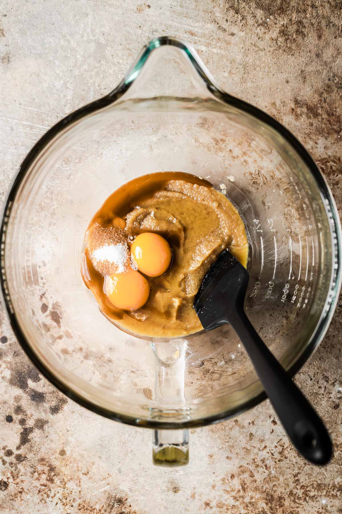 A clear glass mixing bowl with eggs, vanilla and salt being mixed into butter and sugar with a black spatula.