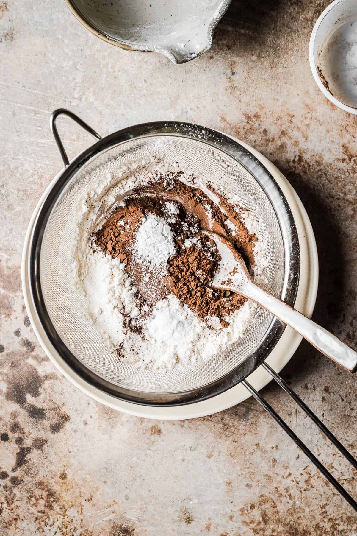 Cocoa powder, flour and baking soda in a sieve over a tan bowl.