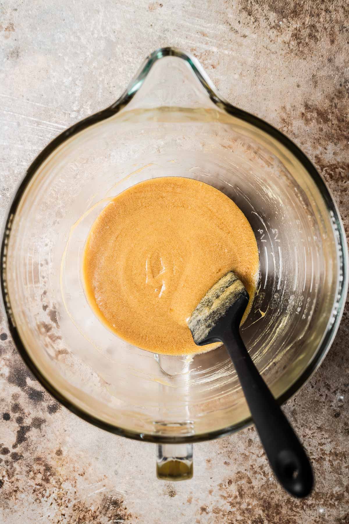 A clear glass mixing bowl with a black spatula with liquid ingredients for chocolate cookies stirred together.