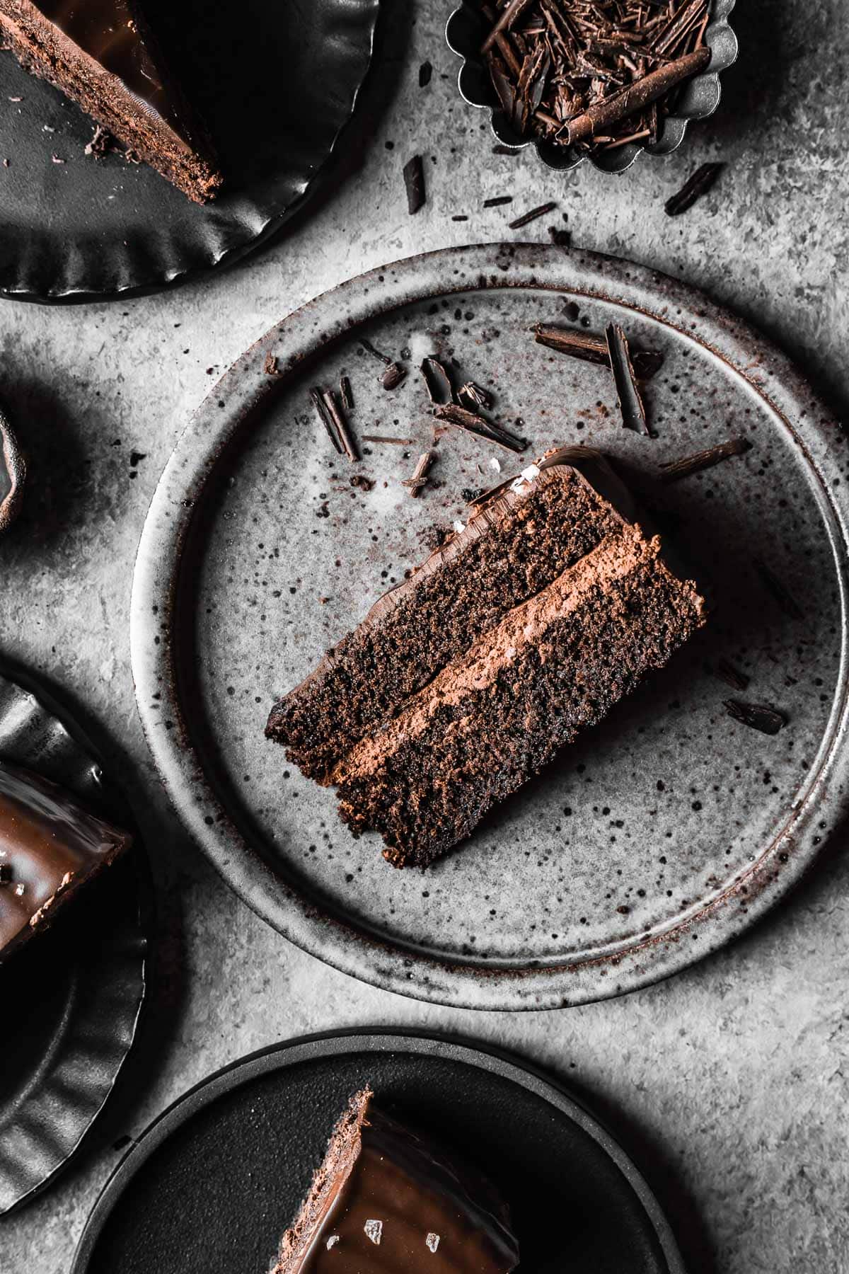 A slice of chocolate cake lying on its side on a speckled grey ceramic plate. There are other plates with cake slices peeking into the frame at left.