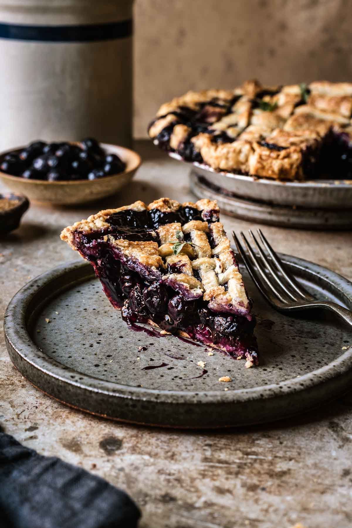 A slice of blueberry pie with a lattice top on a grey speckled ceramic plate. The blueberry filling is well set and bright purple.