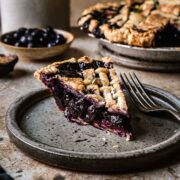 A slice of blueberry pie with a lattice top on a grey speckled ceramic plate. The blueberry filling is well set and bright purple.