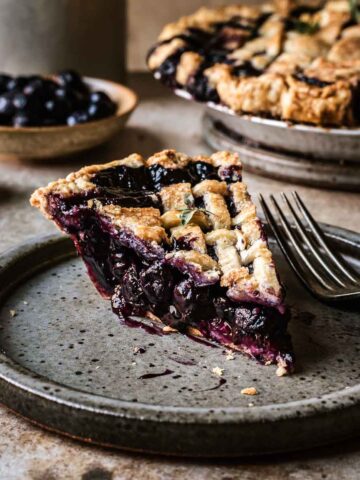 A slice of blueberry pie with a lattice top on a grey speckled ceramic plate. The blueberry filling is well set and bright purple.