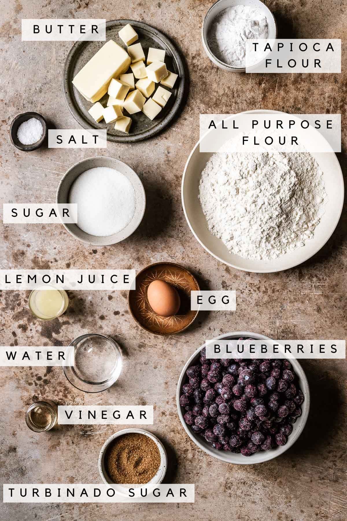 Labeled ingredients in bowls for blueberry pie recipe with frozen blueberries.